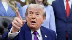 青系のスーツを着たトランプ氏がいすに座り、右手を上げて人差し指で何かを指しながら口を開いている。背後にはスーツ姿の人たちが立っている