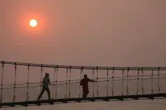 काठमाण्डू उपत्यकामा नाक छोपेर झोलुङ्गेपुल तर्दै गरेका एक व्यक्ति र अर्की महिला पृष्ठभूमिमा धूमिल आकाशमा रातो घाम देखिन्छ