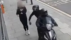 A CCTV image showing a black moped being driven along a pavement by two people wearing all black, snatching a mobile phone from a pedestrian walking along the pavement. One of the people on the moped can be seen holding the phone in their hand moments after snatching it, as the pedestrian recoils.