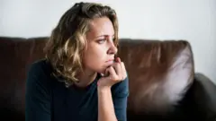 Mulher sentada em sofá com olhar distante apoiando a mão no queixo