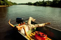 A imagem mostra uma pessoa deitada de forma descontraída em um pequeno barco de alumínio, navegando por um rio largo cercado por densa vegetação. Com um dos braços levantado e o outro apoiado sobre o peito, ela parece apreciar a paisagem, enquanto o motor de popa e um galão de combustível reforçam o clima de expedição pela floresta.