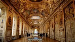 A imagem mostra uma vista geral da galeria Apollon Wing no Louvre, uma sala altamente ornamentada e dourada, com um teto abobadado embelezado e tapeçarias, que abriga as joias da coroa francesa