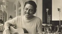 Chico Buarque sorrindo ao tocar violão; ele está aparentemente em um restaurante ou no cômodo ensolarado de uma casa