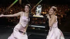 No palco, de costas para uma plateia lotada, Tipnaree "Namtan" Weerawatnodom e Rachanun "Film" Mahawan, com vestidos rosa combinando, uma delas segurando um microfone, sorriem para a câmera.