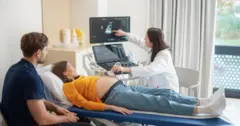 Una mujer embarazada está recostada en una camilla mientras una doctora le toma una imagen del vientre con ultrasonido. Las acompaña la pareja de la mujer, un hombre joven de barba. Todos miran la pantalla que muestra la imagen de ultrasonido
