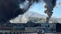 Вид на промзону, над которой в небо поднимаются клубы черного дыма. На заднем плане горы и лес