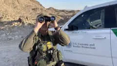 Agente de la patrulla fronteriza usando binoculares junto a su vehículo en una zona desértica de El Paso el 5 de noviembre de 2025.