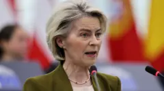 Kobieta o blond włosach, ubrana w oliwkowozieloną kurtkę i naszyjnik, mówi do dwóch mikrofonów