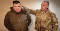 Малюк у кутрці кольору хакі тримає руку на шиї чоловіка у коричневій куртці. У чоловіка руки за спиною 