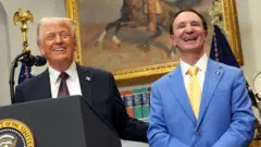 O presidente americano Donald Trump de terno preto e o governador da Louisiana, Jeff Landry, de terno azul. Ambos sorriem em frente a um pódio