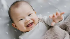 Um bebê sorri e estende a mão em direção à câmera enquanto está deitado com uma manta macia sobre o peito.