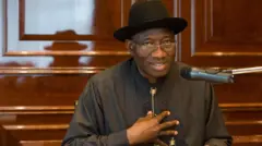Ọmọwe Goodluck Jonathan, aarẹ Naijiria tẹlẹ, o de fila akẹtẹ sori, o logo soju rẹ, o wọsọ alawọ eeru, o si fi ọwọ rẹ si aya.