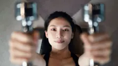 A woman holds weights handles on either side of her face, with the handles out of focus clpose to camera and her face in perfect focus