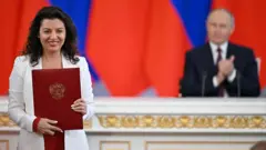 A editora-chefe da RT, Margarita Simonyan, comparece a uma cerimônia de assinatura de documentos. Ao fundo, o presidente russo, Vladimir Putin