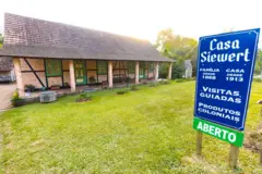 Em frente a casa de tijolinhos, placa diz: 'Casa Siewert, família desde 1868, casa desde 1913, visitas guiadas, produtos coloniais'