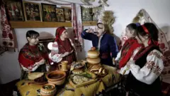 За святковим столом у національному вбранні сидять чоловік, дружина та три дівчини. На стінах ікони та рушники