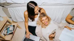 Widok z góry na uśmiechniętą kobietę, która pokazuje smartfon koleżance. Siedzą przy łóżku w akademiku - zdjęcie stockowe