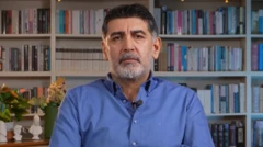 Gültekin bir çalışma odasında kamera karşısında mavi gömleğiyle oturuyor arkasında kitaplık ve kitaplar var. 