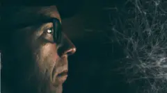 කළු පැහැති කන්නාඩියක් පැළඳි මිනිසෙකුගේ මුහුණ අඳුරු පරිසරයක පෙනේ.