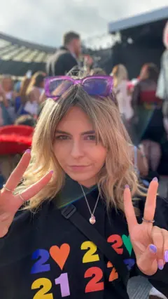 Isla Sneddon posa para a câmera, fazendo sinais de paz. Ela tem cabelo loiro, óculos de sol roxos na cabeça e veste uma blusa preta.