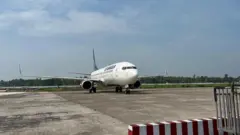 রানওয়েতে দাঁড়িয়ে আছে একটি বিমান, পেছনে দূরে সবুজ গাছপালা দেখা যাচ্ছে