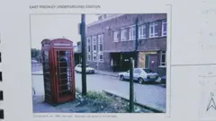 Fotografia colorida de 1984 mostrando uma tradicional cabine telefônica vermelha em uma esquina, com carros estacionados e prédios baixos ao fundo.