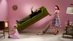 Imagem ilustrativa de uma dona de casa dos anos 1950 limpando a sala com aspirador de pó debaixo do sofá onde um homem está deitado lendo um livro e perto de uma criança sentada assistindo televisão.