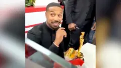 Michael B Jordan smiles at an In-N-Out Burger restaurant as he holds a burger. His Oscar sits on the table next to him.