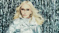 Madonna pozuje w jasnej srebrnej kurtce na tle połyskujących srebrnych pasm.