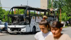 Две женщины идут по улице, за ними — сгоревший автобус.