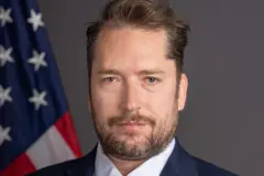 Retrato de Darren Beattie, que aparece sério em frente a bandeira dos EUA