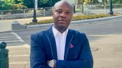  Dr Yves Kaduli, w'imyaka 38 usaba ubuhungiro, ukomoka muri Repubulika ya Demokarasi ya Congo, arahagaze asobekeranyije amaboko mu gatuza yambaye ikoti ricyeye