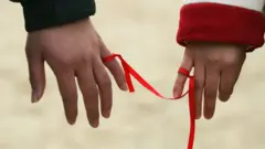 Duas pessoas amarram uma fita vermelha nos dedos mindinhos