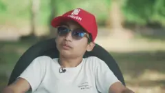 João do Clima é uma criança. Ele está sentado em uma cadeira e usa óculos escuros e um boné vermelho