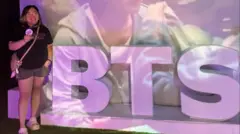 Cookie posa ao lado de uma sinalização roxa do BTS em um evento organizado pelo Exército