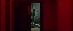 Pasillo iluminado en rojo con una puerta abierta que muestra una sala con casilleros y papeles esparcidos en el suelo