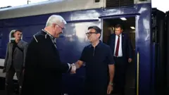 Burnham îi strânge mâna lui Sybiha, în timp ce amândoi zâmbesc, stând alături de alți doi bărbați și lângă un tren.