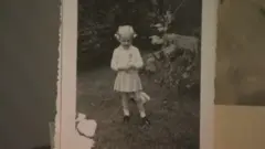 Una antigua fotografía en blanco y negro de Lana Ponting cuando era niña. Lleva un vestido de color claro y su pelo rubio corto está recogido en dos coletas.