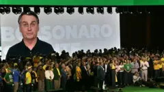 Imagem de Jair Bolsonaro no telão atrás do palco.