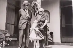 Alberto Veraza, vestido com terno, gravata e chapéu, está com as mãos para trás. Ao lado dele está Adriana, que tem cabelos brancos, usa óculos e um vestido escuro. Bem perto dela está Mara, que tem tranças, veste um vestido branco e segura um cachorro. Os três olham para a câmera e estão no que parece ser o pátio de uma casa