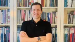 O professor Raúl Cordero, um homem branco, veste uma camisa preta de mangas curtas e está em frente a uma estante de livros.