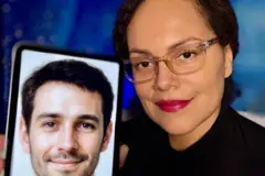 Uma mulher de óculos transparentes e batom vermelho vivo sorri enquanto segura o celular em frente à câmera, mostrando uma foto de seu assistente virtual. Na tela do celular, há a foto de um homem com cabelo curto, barba rala e olhos sorridentes.