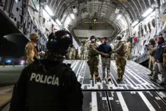 Двое военных с закрытыми масками лицами ведут мужчину по трапу, его встречает полицейский.