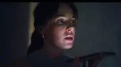 Uma mulher segurando um smartphone perto da boca, vestindo uma blusa cinza sem mangas, em um ambiente interno com pouca luz.