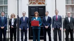 スーツを着た男女が7人、屋外に横並びで立っている。中央の青いパンツスーツの女性が、赤い箱型のかばんを胸元に掲げている。背後には白い建物と黒い鉄柵、黒い玄関のドアが写っている