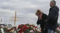 Dos personas lloran frente a un montón de flores. 