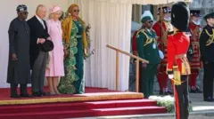 Aarẹ Bola Tinubu to wọ asọ buba ati sokoto alawọ dudu pẹlu fila ati igo loju, duro sẹgbẹ ọba Charles to wọ kootu dudu, o mu fila lọwọ, ti olori Camilla to wọ asọ alawọ pinki ati ate lori si duro ti ọba nigba ti aya aarẹ Naijiria, Oluremi Tinubu duro sikẹrin wọn, o wọ kaba gbagẹrẹ alawọ eweko, o we gele asọ oke alawọ goolu, to si da ipele asọ oke naa bo ọrun rẹ. n ẹsọ ọba ilẹ Gẹẹsi to sọwọ ọtun wọn.