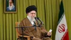 Hamaney bir koltukta oturuyor. Önünde mikrofon. Arkasında İran bayrağı ve Humeyni'nin fotoğrafı var.