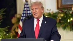 ダークスーツ姿のトランプ氏が演台で話している。背後には米国旗やクリスマスっぽい装飾がぼんやり見える