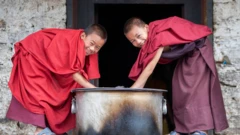 Dos jóvenes monjes budistas agachados limpiando una gran olla. Miran sonrientes a la cámara. 